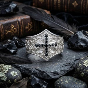 Könnte beinhalten: Ein silbernes Armreif-Armband mit einem verzierten Blumen- und Scroll-Design. Ein schwarzes Kreuz ist auf dem Armband zentriert, mit schwarzen Steinen im Kreuz. Das Armband wird auf einer dunklen, strukturierten Oberfläche präsentiert.