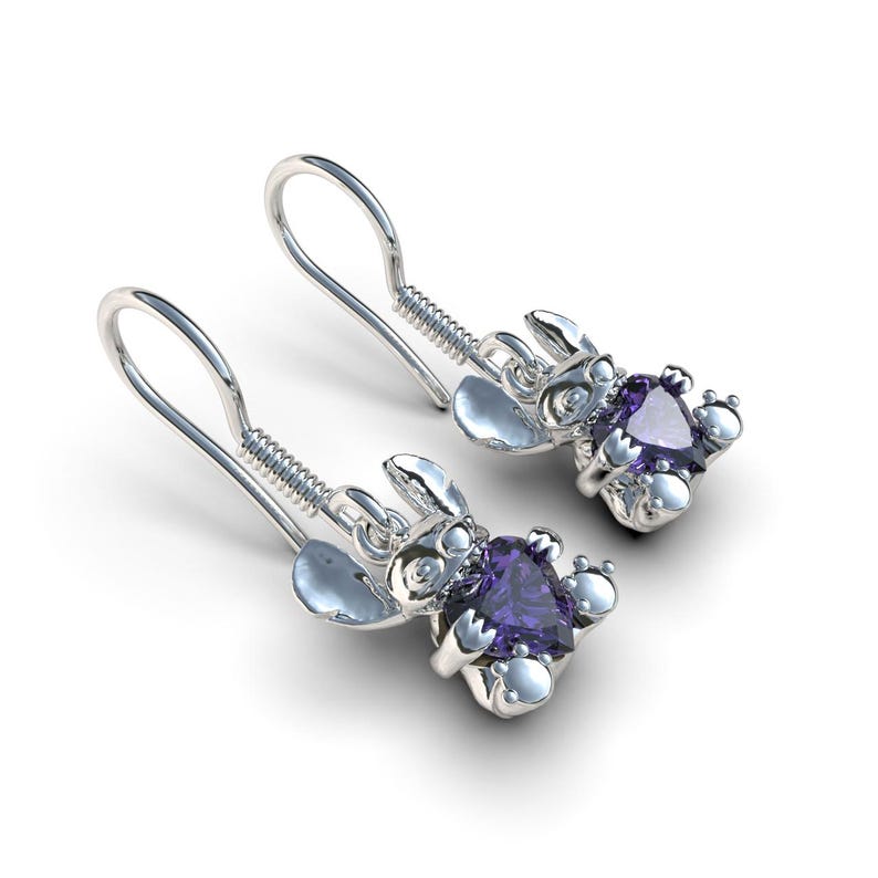 May include: A pair of silver-colored earrings featuring a cartoon character design. Each earring has a purple gemstone at the center of the character's body. The earrings have a hook style closure.