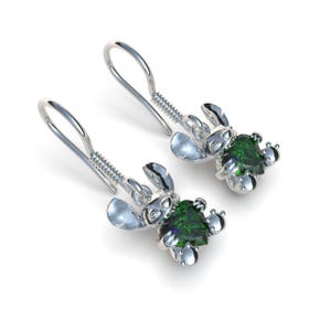 May include: A pair of silver-colored dangle earrings featuring a cartoon character design. Each earring has a green heart-shaped gemstone at the center. The earrings have a hook closure and are set against a white background.