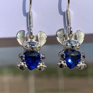 May include: A pair of silver-toned earrings featuring a cartoon character design. Each earring has a small figure with large ears and a heart-shaped, deep blue gemstone. The earrings hang from a silver hook with a coiled accent.