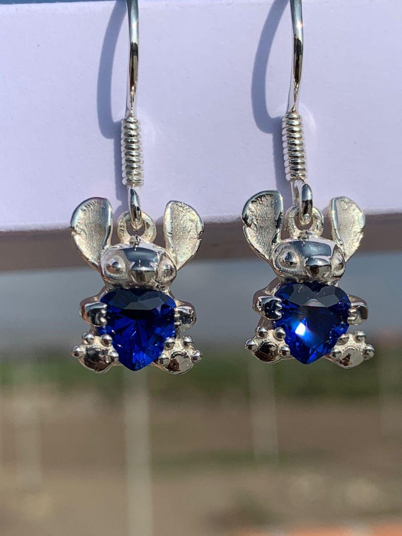 May include: A pair of silver-toned earrings featuring a cartoon character design. Each earring has a small figure with large ears and a heart-shaped, deep blue gemstone. The earrings hang from a silver hook with a coiled accent.