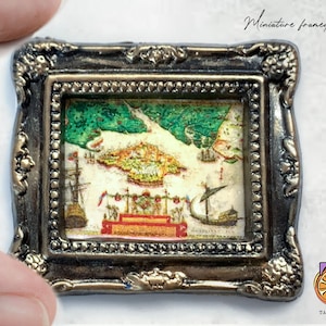 May include: A miniature framed art piece with a vintage map of a city with ships and a red and gold building in the center. The frame is ornate and has a gold finish.