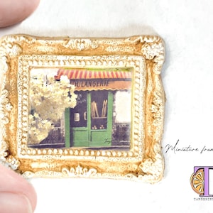 May include: A miniature framed art print of a French bakery with a green door and a sign that reads "Boulangerie". The frame is a light brown color with a white ornate design.