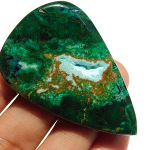 Puede incluir: Una piedra preciosa pulida con una vibrante paleta de colores verdes y turquesas. La piedra presenta una cavidad central con acentos blancos y marrones. La gema se sostiene en una mano, mostrando su forma única y patrones naturales. Un hermoso ejemplo de piedra natural.