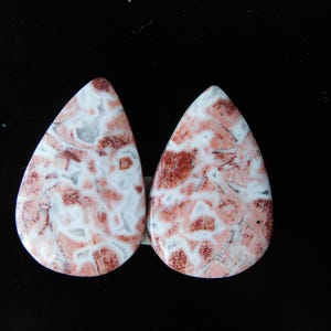 Könnte beinhalten: Zwei tropfenförmige Edelsteine mit weißer Basis und einem Muster aus roten und grauen Flecken. Die Steine sind vor einem schwarzen Hintergrund platziert, was ihre einzigartigen Farb- und Musterunterschiede hervorhebt. Ideal für die Schmuckherstellung.