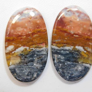Op de afbeelding: Een paar ovale cabochon edelstenen met een gelaagd patroon van witte, oranje, bruine en grijze kleuren.