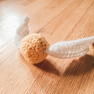 May include: A crocheted golden snitch with white wings. The snitch is a popular character from the Harry Potter series.