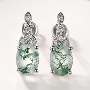 May include: A pair of silver-toned stud earrings. Each earring features an oval-shaped, light green stone with dark green veining. The top of each earring has a Celtic knot design with small, clear stones.