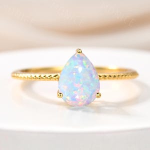 May include: A gold ring featuring a pear-shaped opal gemstone. The opal displays a mix of blue, white, and pink hues. The ring band has a textured design, and the opal is held in place by four small prongs.