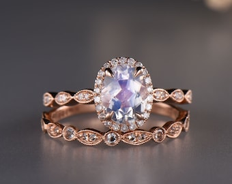 May include: A rose gold engagement ring set with a large oval moonstone center stone surrounded by a halo of smaller diamonds. The matching wedding band features a delicate design with small diamonds.