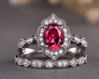 May include: A vintage-inspired engagement ring set featuring a large oval-shaped ruby surrounded by a halo of smaller diamonds. The band is adorned with delicate diamond accents and a matching wedding band.