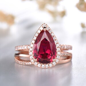 Op de afbeelding: Een peervormige robijn verlovingsring gezet in roségoud met een halo van kleinere diamanten. De ring heeft ook een delicate, drievoudige diamant trouwband.
