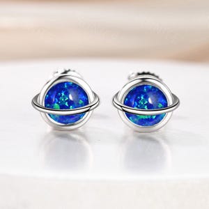 May include: A pair of silver stud earrings. Each earring features a blue opal stone in a planet design, with a silver band encircling the stone. The opal has a speckled appearance with shades of blue and green.