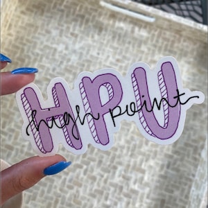 K&ouml;nnte beinhalten: Ein Aufkleber mit dem Text "HPU High Point" in einem lila-wei&szlig;en Design. Die Buchstaben "HPU" sind fett umrandet, w&auml;hrend "High Point" in Schreibschrift gehalten ist. Der Aufkleber befindet sich vor einem strukturierten Hintergrund.