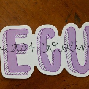 Puede incluir: Una pegatina blanca con el texto "ECU East Carolina" en letras moradas con burbujas. Las letras tienen un efecto 3D con l&iacute;neas diagonales para sombrear. Las palabras est&aacute;n escritas a mano.