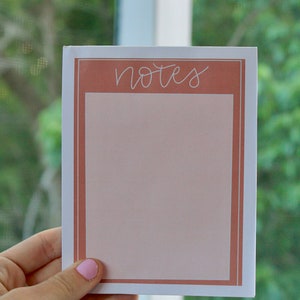 May include: A notepad with a white border and a peach-colored interior. The word "notes" is written in cursive at the top. The notepad is held in a hand with a pink painted nail.