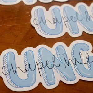 May include: Several stickers with the text "UNC Chapel Hill" in a light blue bubble letter style with a black outline. The stickers are on a wooden surface.