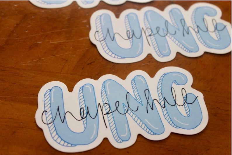 May include: Several stickers with the text "UNC Chapel Hill" in a light blue bubble letter style with a black outline. The stickers are on a wooden surface.