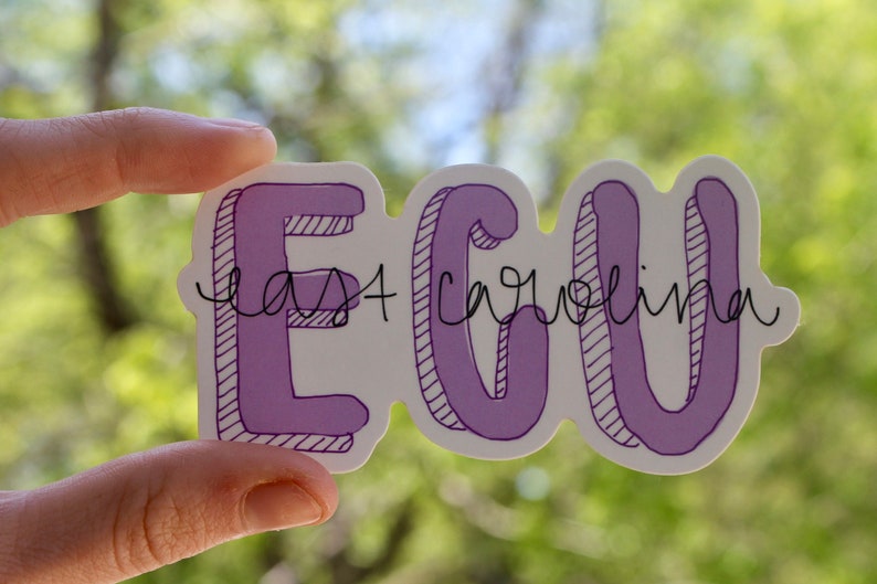 Puede incluir: Una pegatina con la palabra "ECU" en morado, delineada en negro, con las palabras "East Carolina" en escritura cursiva. La pegatina se sostiene contra un fondo verde borroso.
