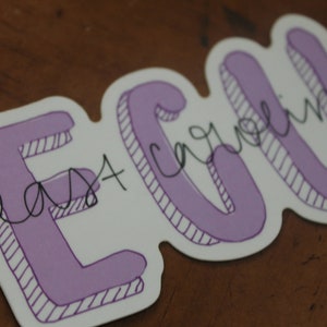 Puede incluir: Una pegatina con las letras "ECU" en una fuente morada con contorno. Las palabras "East Carolina" est&aacute;n escritas en cursiva sobre las letras. La pegatina tiene un borde blanco y est&aacute; sobre una superficie marr&oacute;n.