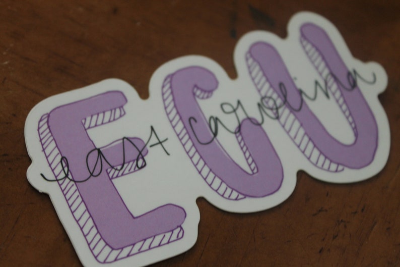 Puede incluir: Una pegatina con las letras "ECU" en una fuente morada con contorno. Las palabras "East Carolina" est&aacute;n escritas en cursiva sobre las letras. La pegatina tiene un borde blanco y est&aacute; sobre una superficie marr&oacute;n.