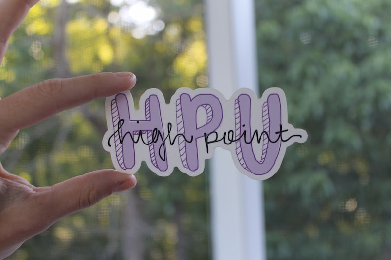 K&ouml;nnte beinhalten: Ein Aufkleber mit den Buchstaben "HPU" in hellem Lila, mit den Worten "high point" in Schreibschrift. Der Aufkleber hat einen wei&szlig;en Rand und wird vor einem verschwommenen gr&uuml;nen Hintergrund gehalten.
