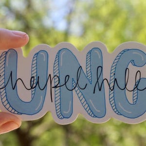 Könnte beinhalten: Ein Aufkleber mit dem Text "UNC chapel hill". Die Buchstaben "UNC" sind hellblau mit weißem Rand, und die Worte "chapel hill" sind in schwarzer Schreibschrift. Der Aufkleber wird von einer Hand gehalten.