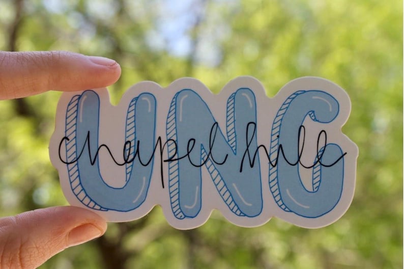 May include: A sticker with the text "UNC chapel hill" in a bubble letter style. The letters "UNC" are light blue with a white outline, and the words "chapel hill" are in a black cursive font. The sticker is held by a hand.