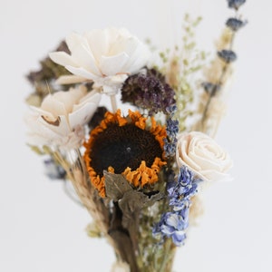 May include: A dried flower arrangement in a clear glass jar. The arrangement includes white, blue, and orange flowers, as well as dried grasses.
