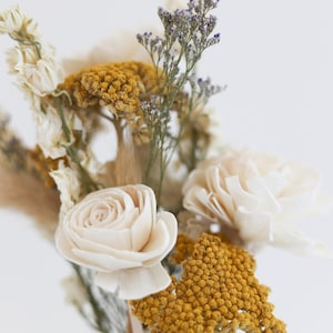 May include: A bouquet of dried flowers in a clear glass vase. The bouquet includes white and cream colored flowers, yellow flowers, and purple flowers.