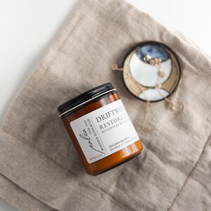 May include: A brown glass jar with a black lid containing Driftwood Restorative Botanical Body Butter for the Modern Bohemian. The jar is sitting on a beige linen fabric with a gold chain necklace and a ceramic dish with a blue and white design.