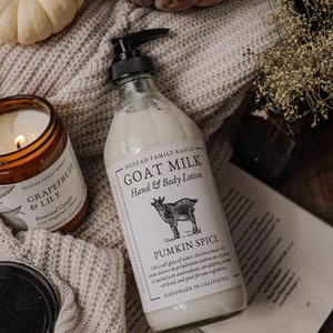 Może przedstawiać: Szklana butelka z mleczkiem kozim do rąk i ciała z białą etykietą, na kt&oacute;rej widnieje napis "Goat Milk Hand & Body Lotion" i "Pumpkin Spice". Butelka stoi na dzianinowym kocu z zapalonym świecą i dyniami w tle.