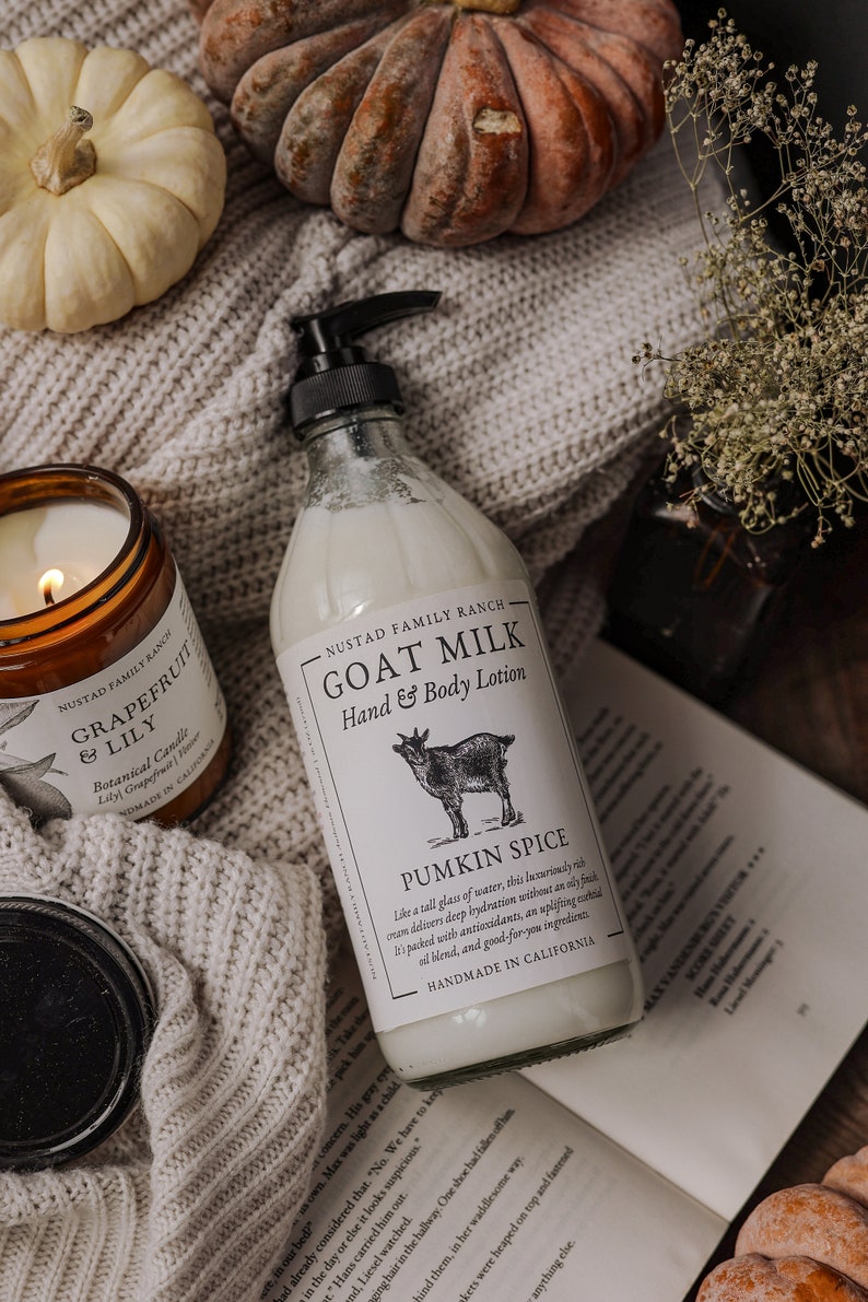 Może przedstawiać: Szklana butelka z mleczkiem kozim do rąk i ciała z białą etykietą, na kt&oacute;rej widnieje napis "Goat Milk Hand & Body Lotion" i "Pumpkin Spice". Butelka stoi na dzianinowym kocu z zapalonym świecą i dyniami w tle.