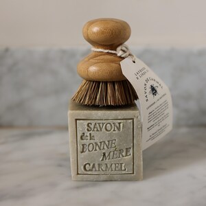 Puede incluir: Una pastilla de jab&oacute;n rectangular con las palabras "SAVON de la BONNE MERE CARMEL" en relieve, coronada por un cepillo de madera. Una etiqueta est&aacute; unida al cepillo con las palabras "LAVENDER & LINEN CO" y una ilustraci&oacute;n de abeja.