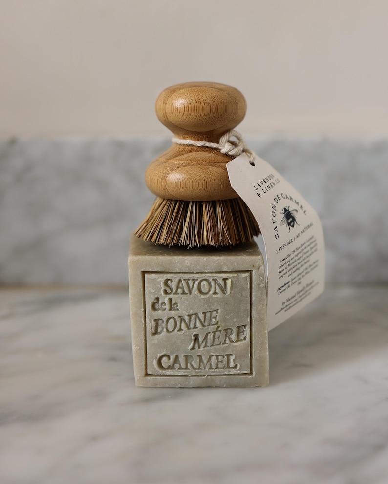 Puede incluir: Una pastilla de jab&oacute;n rectangular con las palabras "SAVON de la BONNE MERE CARMEL" en relieve, coronada por un cepillo de madera. Una etiqueta est&aacute; unida al cepillo con las palabras "LAVENDER & LINEN CO" y una ilustraci&oacute;n de abeja.