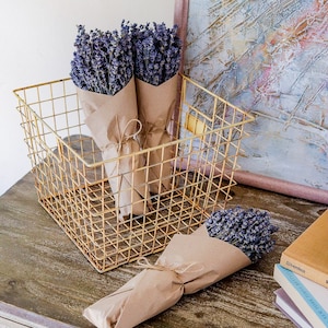 May include: Three bundles of dried lavender flowers wrapped in brown paper are displayed in a gold wire basket. The basket is sitting on a wooden surface.