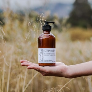 Może przedstawiać: Brązowa butelka ze szkła z czarną pompką, zawierająca balsam do ciała. Butelka jest oznaczona etykietą "New Dawn Replenishing Botanical Body Lotion" i "For the Modern Bohemian".