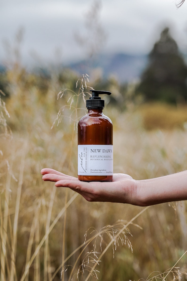 Może przedstawiać: Brązowa butelka ze szkła z czarną pompką, zawierająca balsam do ciała. Butelka jest oznaczona etykietą "New Dawn Replenishing Botanical Body Lotion" i "For the Modern Bohemian".