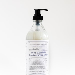 May include: A clear glass bottle with a black pump dispenser filled with a white liquid. The bottle is labeled "NUSTAD FAMILY RANCH HAND CRAFTED | SMALL BATCH" and "PURE CASTILLE HAND & BODY SOAP" with the scent "lavender". The label also states "Authentic Elemental | Palm Oil Free | Made in the USA HANDMADE IN CALIFORNIA".