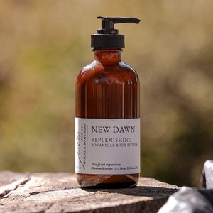 Może przedstawiać: Brązowa szklana butelka z czarną pompką, z etykietą "NEW DAWN REPLENISHING BOTANICAL BODY LOTION". Etykieta zawiera r&oacute;wnież informacje o składnikach: "Czysty składnik roślinny: Ekstrakt z rumianku - Olej z konopi i maruli".