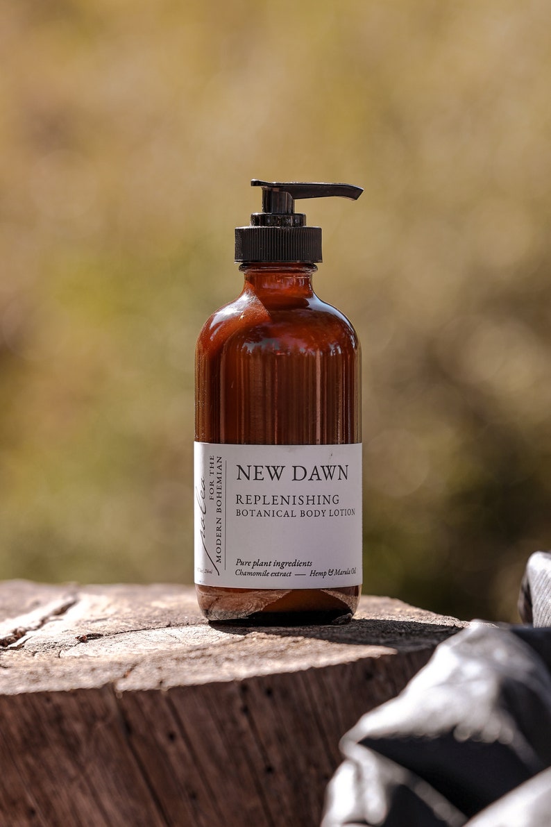 Może przedstawiać: Brązowa szklana butelka z czarną pompką, z etykietą "NEW DAWN REPLENISHING BOTANICAL BODY LOTION". Etykieta zawiera r&oacute;wnież informacje o składnikach: "Czysty składnik roślinny: Ekstrakt z rumianku - Olej z konopi i maruli".