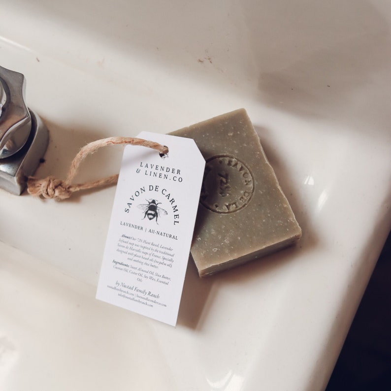 Puede incluir: Una pastilla de jab&oacute;n verde y una etiqueta blanca con el texto "Lavender & Linen Co" y "Savon de Carmel" se muestran. La etiqueta presenta una ilustraci&oacute;n de abeja y las palabras "Lavender All-Natural". El jab&oacute;n est&aacute; sobre una superficie blanca.