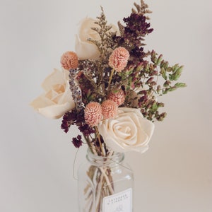 Könnte beinhalten: Eine Glasvase mit einem weißen Etikett mit der Aufschrift "Floral Diffuser" gefüllt mit einem Strauß getrockneter Blumen. Der Strauß enthält weiße Rosen, rosa und violette Wildblumen und getrocknete Grünpflanzen.