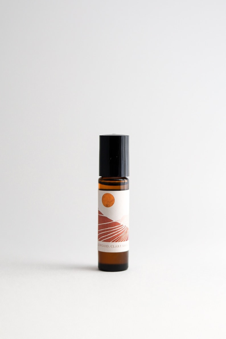 May include: A brown glass roller bottle with a black cap and a white label. The label features a graphic of a sunset over a desert landscape with the text "SANDALWOOD, CLARY SAGE".