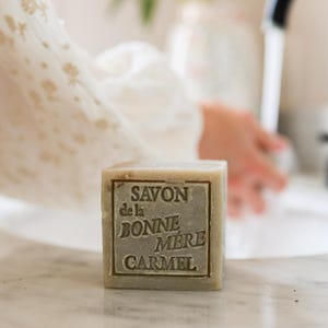 Puede incluir: Una pastilla de jab&oacute;n cuadrada de color verde claro con el texto "SAVON de la BONNE MERE CARMEL" en relieve. El jab&oacute;n est&aacute; sobre una superficie de m&aacute;rmol, con las manos de una persona y un grifo al fondo.