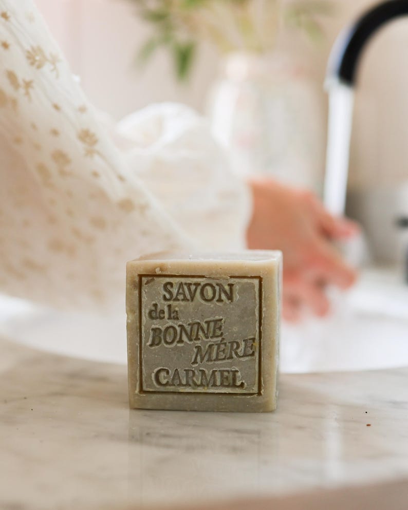 Puede incluir: Una pastilla de jab&oacute;n cuadrada de color verde claro con el texto "SAVON de la BONNE MERE CARMEL" en relieve. El jab&oacute;n est&aacute; sobre una superficie de m&aacute;rmol, con las manos de una persona y un grifo al fondo.