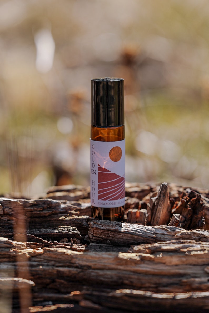 Może przedstawiać: Brązowa szklana butelka z rolką z czarną zakrętką i białą etykietą z napisem "Golden Hour Chamomile Lavender Roll-On". Etykieta zawiera grafikę zachodu słońca nad pasmem g&oacute;rskim.