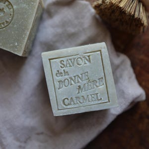 Puede incluir: Primer plano de dos jabones cuadrados de color verde claro con texto en relieve. Un jab&oacute;n muestra el texto "SAVON de la BONNE MERE CARMEL". Los jabones descansan sobre una tela beige, con un cepillo con mango de madera en el fondo. Los jabones parecen hechos a mano.