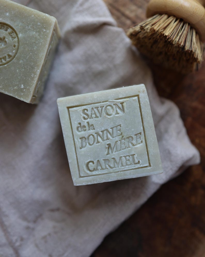 Puede incluir: Primer plano de dos jabones cuadrados de color verde claro con texto en relieve. Un jab&oacute;n muestra el texto "SAVON de la BONNE MERE CARMEL". Los jabones descansan sobre una tela beige, con un cepillo con mango de madera en el fondo. Los jabones parecen hechos a mano.