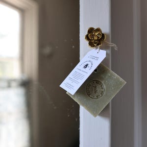 Puede incluir: Un pomo dorado en forma de flor sujeta una etiqueta y una pastilla de jab&oacute;n verde. La etiqueta dice "Lavender & Linen Co." y "Savon de Carmel." El jab&oacute;n tiene un dise&ntilde;o estampado. El fondo es un marco de puerta blanco.